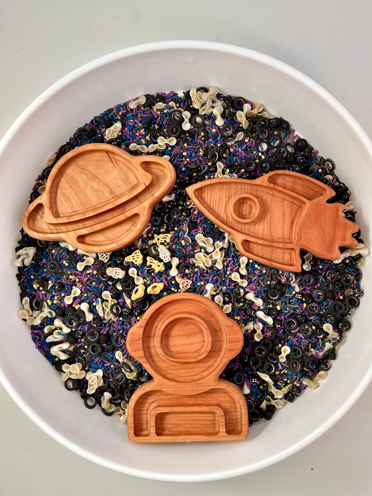 Wooden trays shaped like a rocket, planet with rings, and astronaut on mixed black, purple, and white sensory rice and pasta shapes