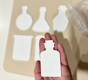 Hand holding a white dry erasable potion bottle shape with five other beaker and bottle shapes on beige board background