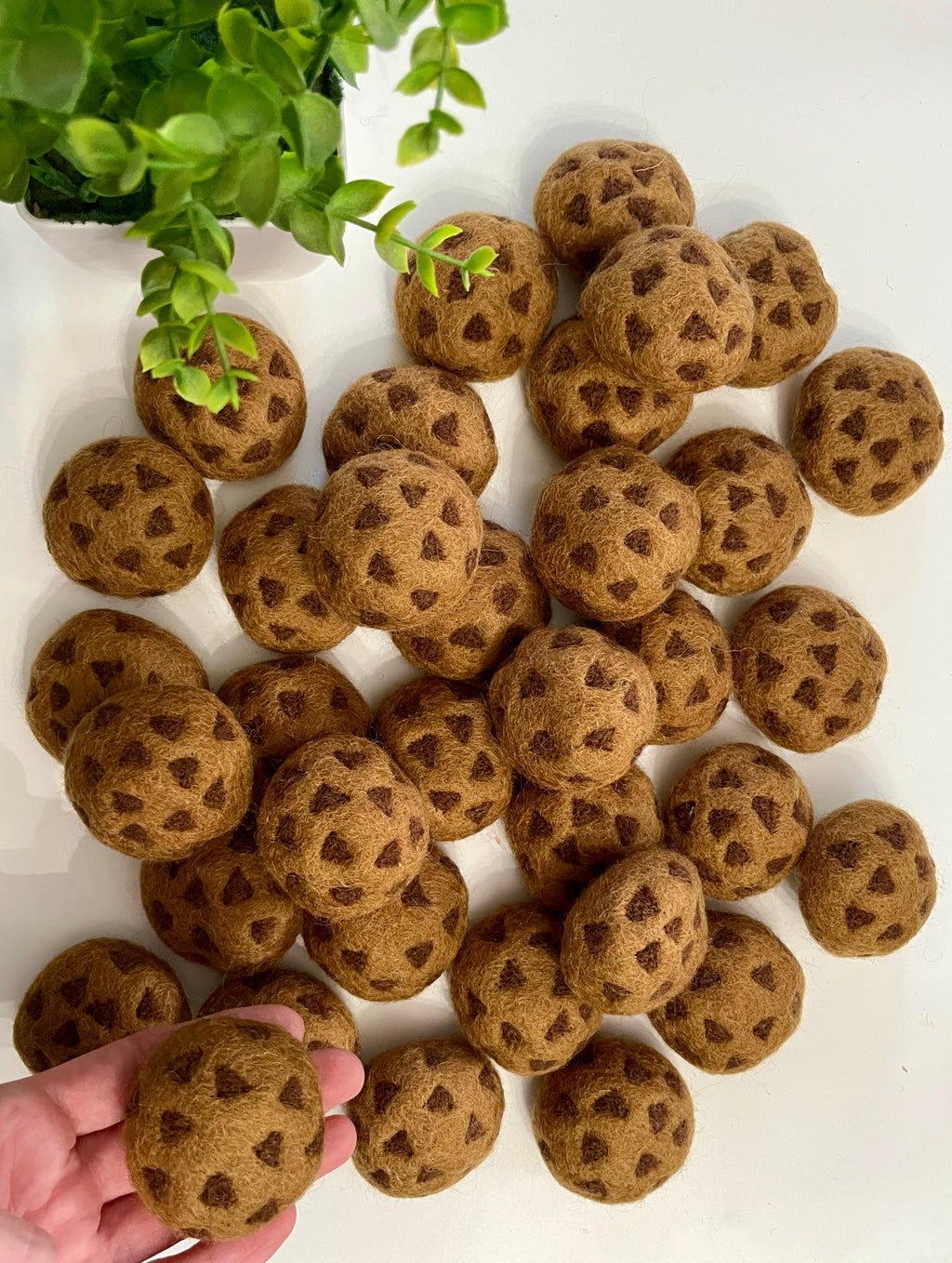 Hand holding a brown felt ball with dark spots, surrounded by similar felt balls on white surface near green plant leaves