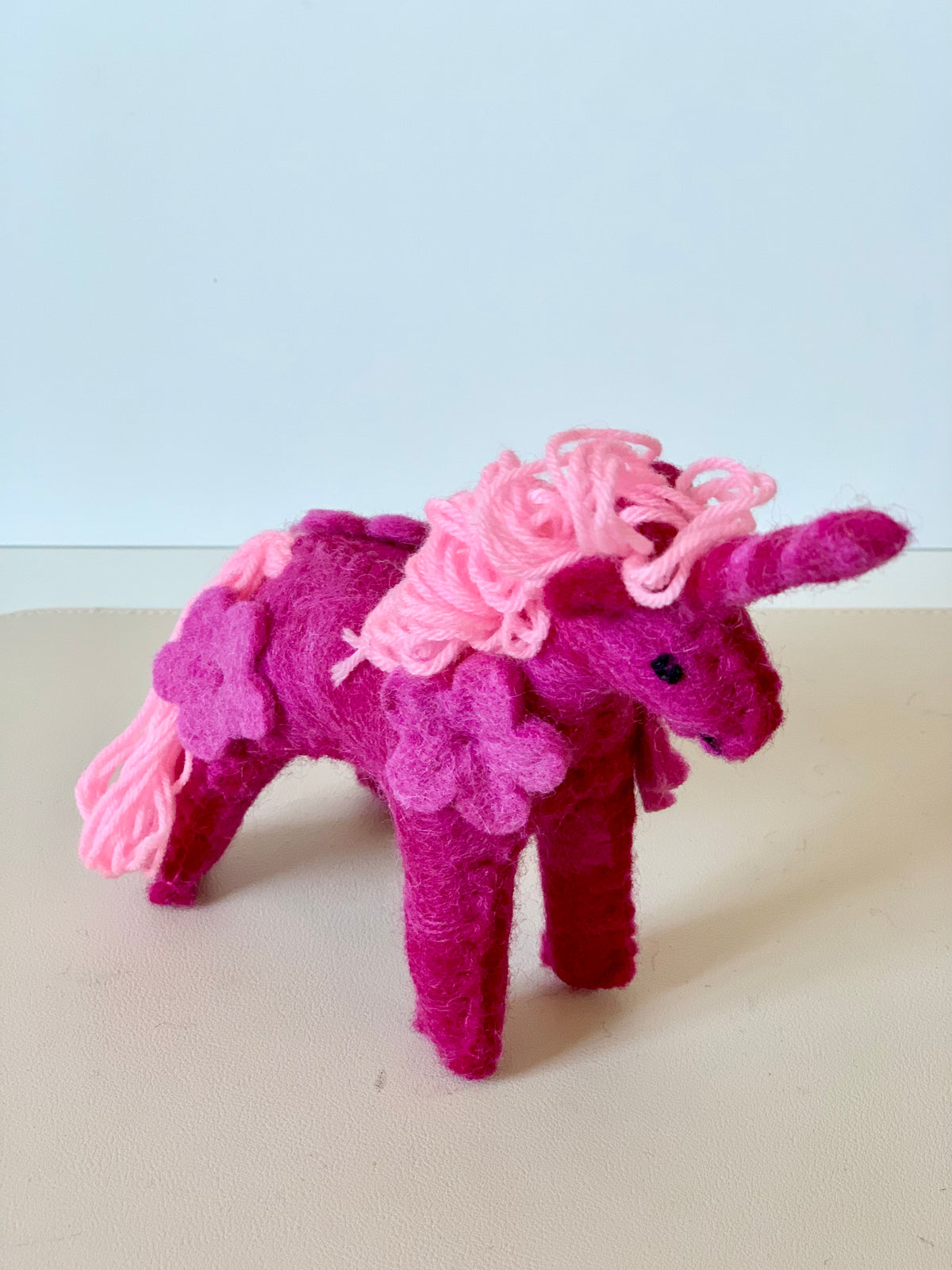 Plush felt unicorn figurine in shades of pink with yarn mane and floral accents standing on a beige surface