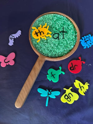 Wooden magnifying glass sensory tray filled with green rice and colorful insect-shaped letter tiles spelling phonics blends on dark background