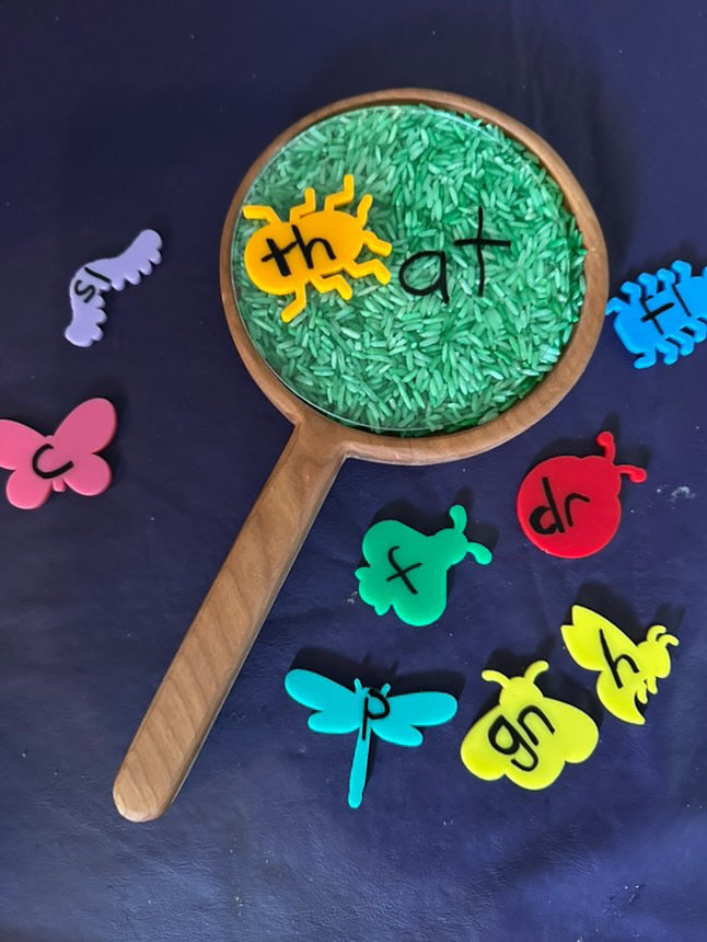 Wooden magnifying glass sensory tray filled with green rice and colorful insect-shaped letter tiles spelling phonics blends on dark background