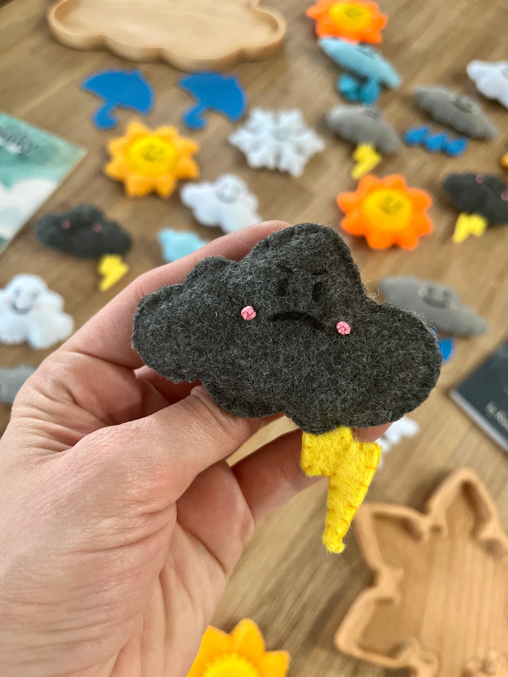 Hand holding a dark gray felt cloud with an angry face and yellow lightning bolt, surrounded by colorful Felt Weather Shapes, Made in Canada, handcrafted, homeschool, education resources.