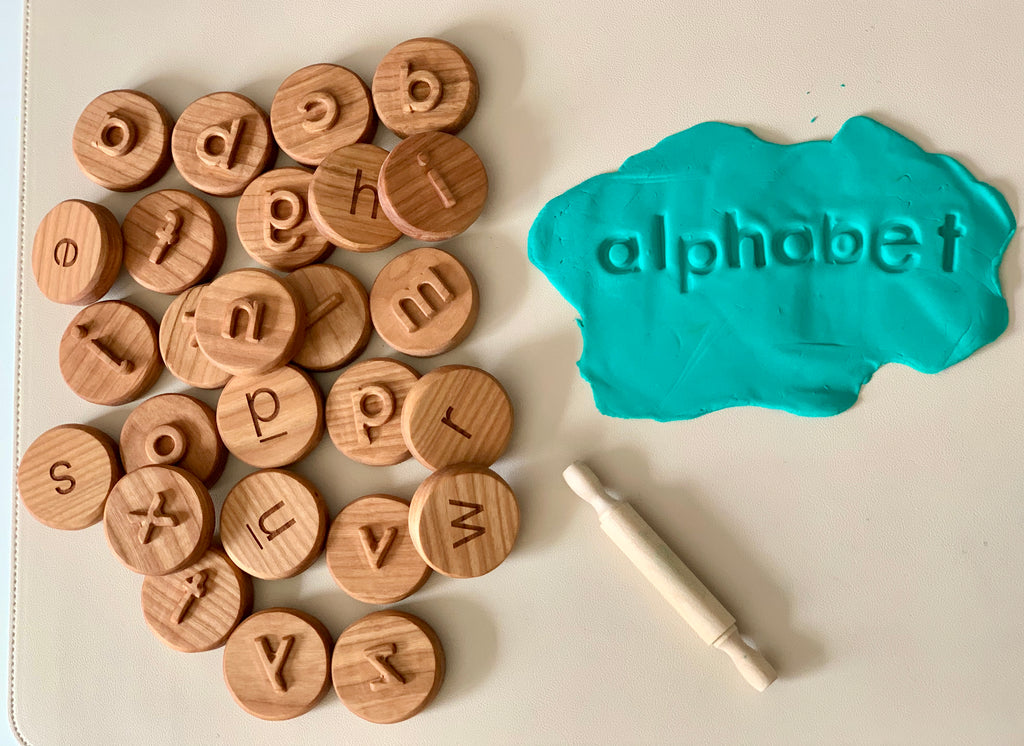Wooden alphabet tokens with lowercase letters next to teal playdough stamped with the word "alphabet" and a small rolling pin