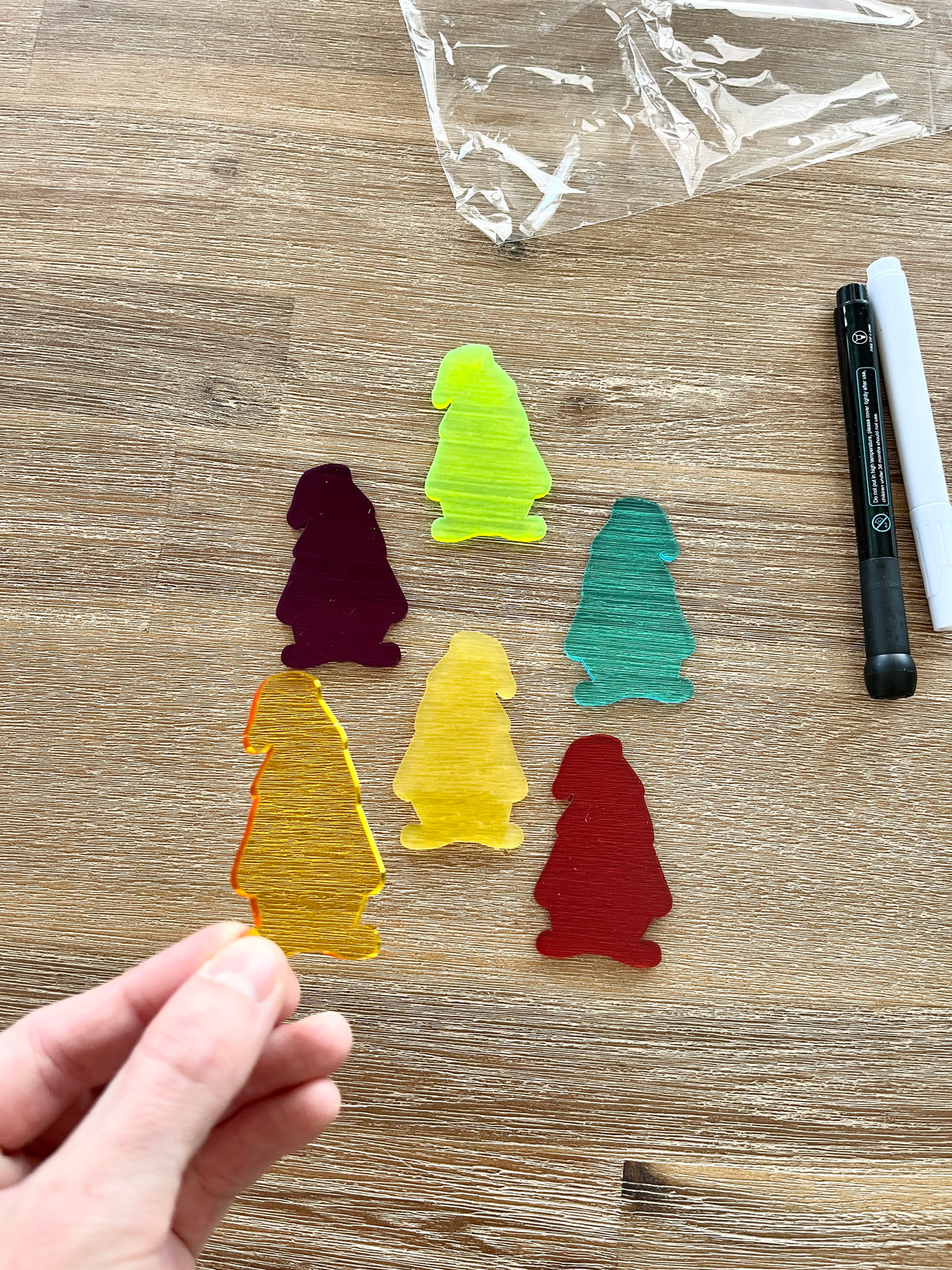 Hand holding a translucent yellow gnome-shaped piece with five additional colorful translucent gnomes and black and white dry erase markers on a wood surface, featuring Little Dry Erasables - Translucent Gnomes, handcrafted homeschool education resources.