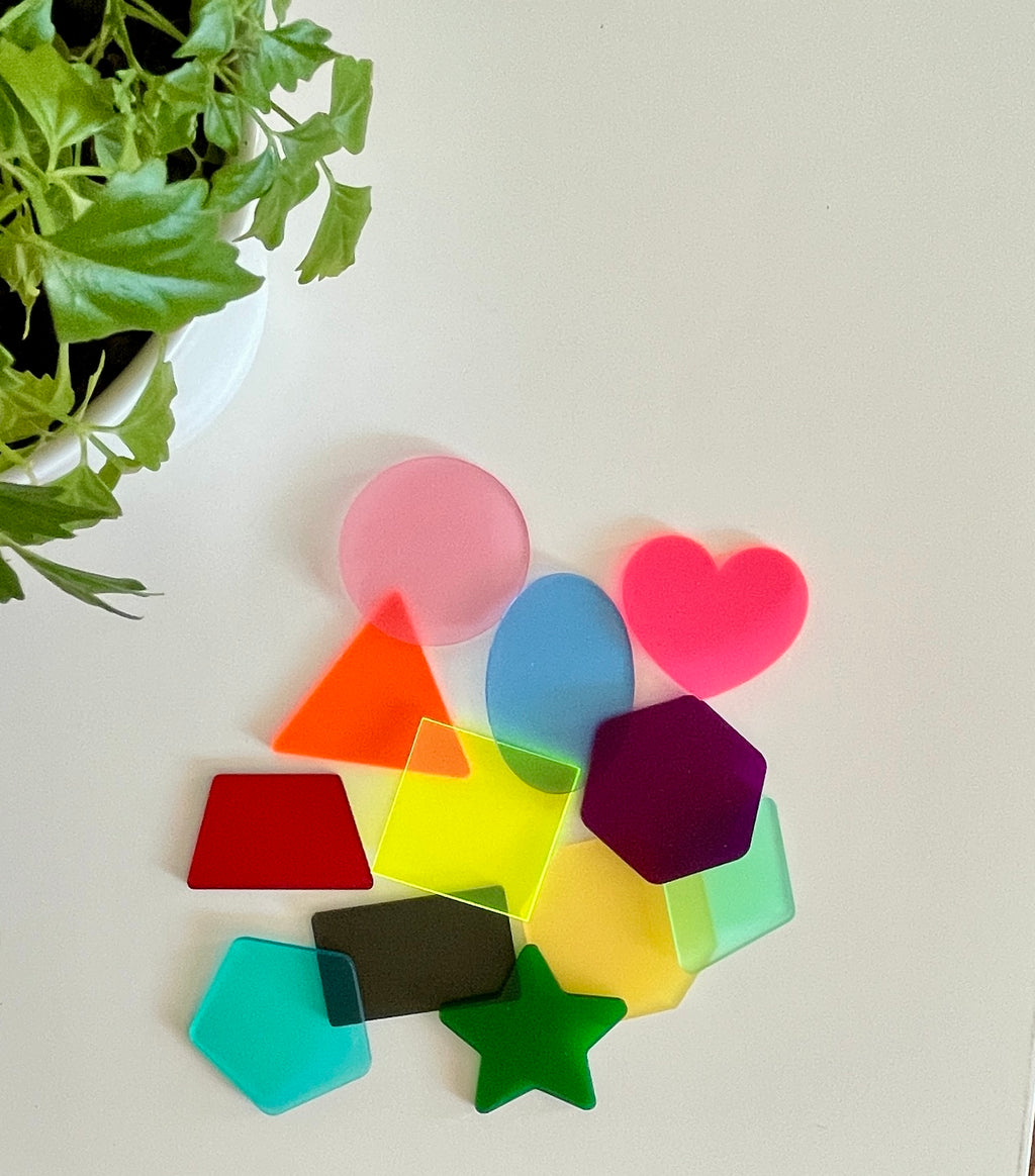 Collection of translucent colorful acrylic shapes including circle, heart, star, and polygons on white surface near green plant