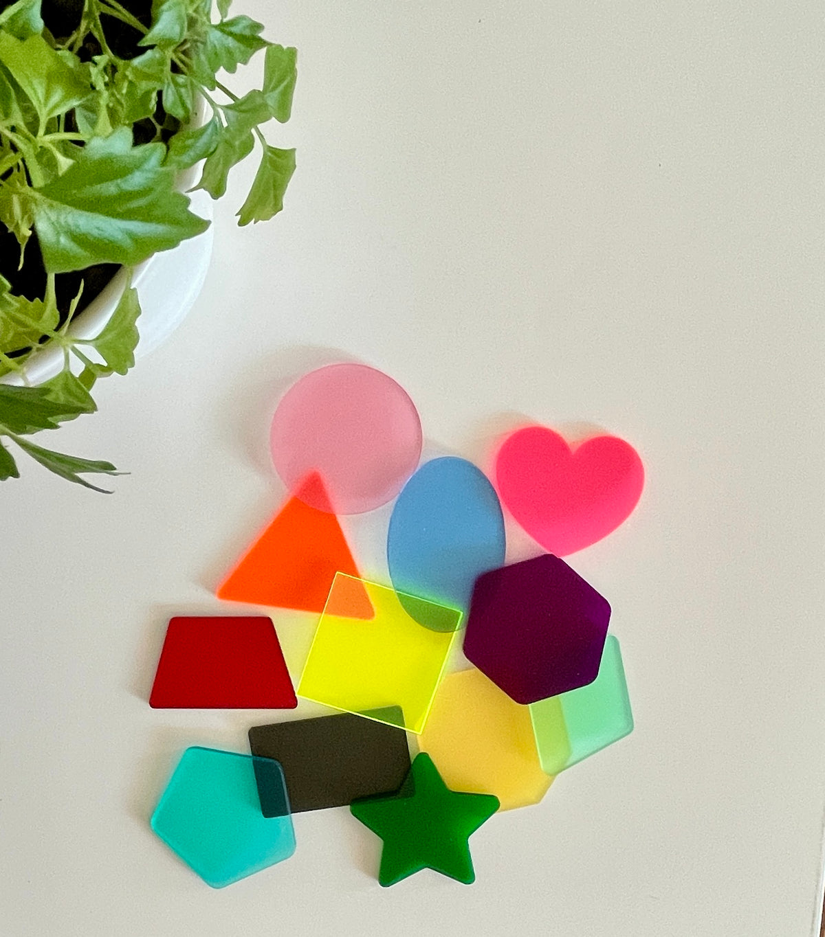 Collection of translucent colorful acrylic shapes including circle, heart, star, and polygons on white surface near green plant