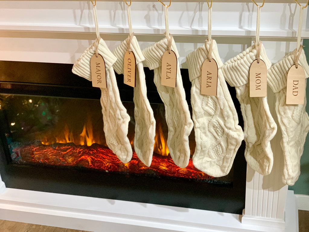 Six cream-colored cable knit Christmas stockings hanging on white mantle above a glowing electric fireplace