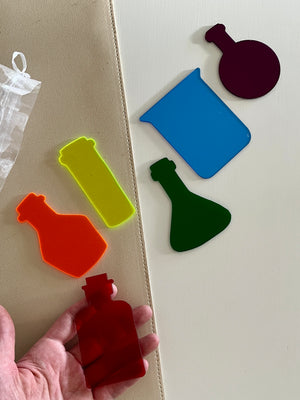 Set of six colorful dry erasable potion bottle and beaker shapes on beige and white surfaces with hand holding one piece
