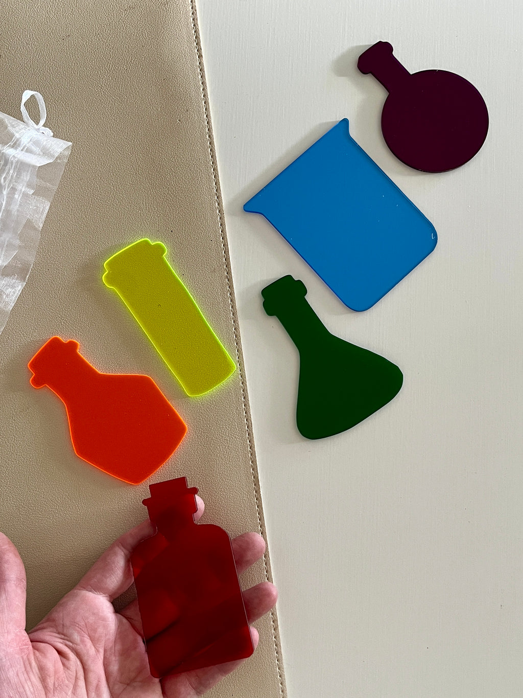 Set of six colorful dry erasable potion bottle and beaker shapes on beige and white surfaces with hand holding one piece