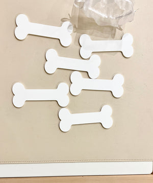 Six plain white bone-shaped tags arranged on a beige surface next to a clear plastic object