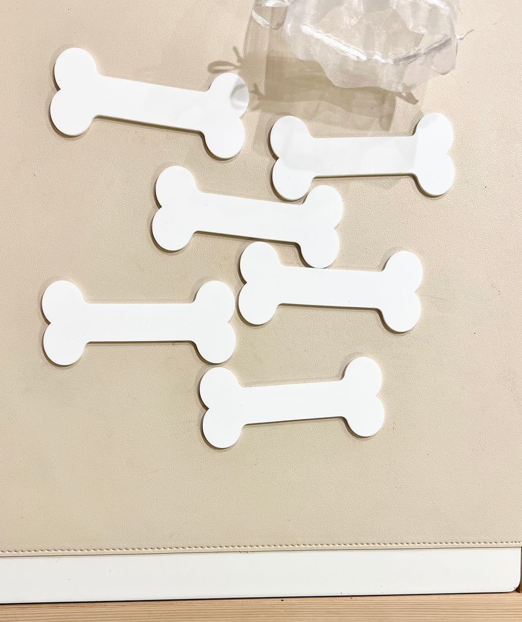Six plain white bone-shaped tags arranged on a beige surface next to a clear plastic object