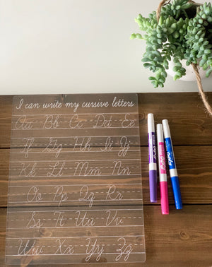 Transparent cursive writing practice board with white letters and three colored dry erase markers on wooden surface near plant