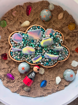 Cloud-shaped wooden sensory tray filled with turquoise beads, colorful shiny shapes, and sand with scattered shells and pirate figure