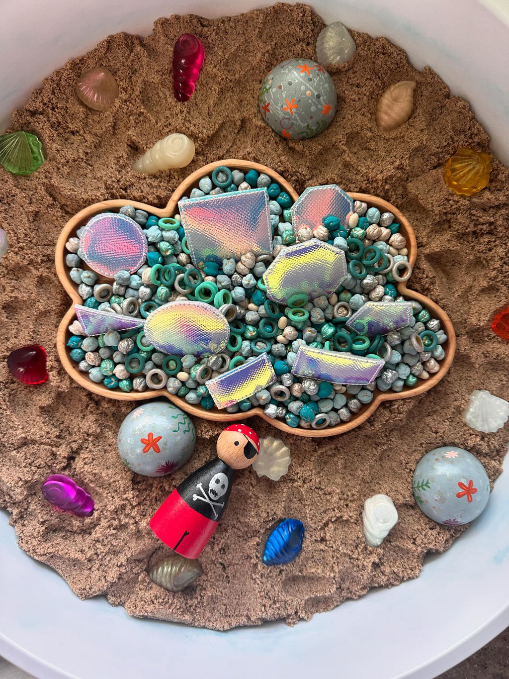 Cloud-shaped wooden sensory tray filled with turquoise beads, colorful shiny shapes, and sand with scattered shells and pirate figure