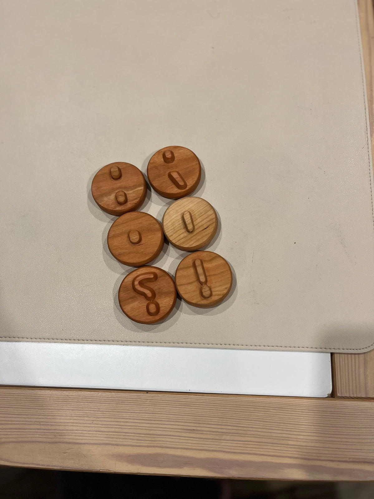 Seven round wooden tokens engraved with various punctuation marks on a light fabric surface above a wooden table edge