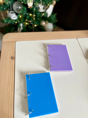 Two small plastic binders with blue and purple covers lying on a white table near a decorated Christmas tree