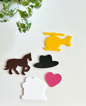 Five colorful acrylic shapes including yellow helicopter, brown horse, black hat, white house, and pink heart on white background with green plant nearby