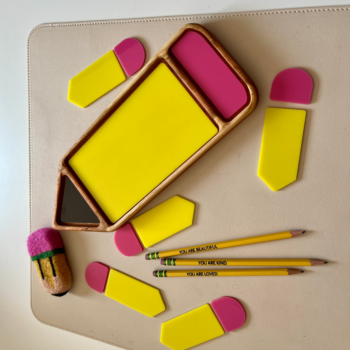 Wooden pencil-shaped tray holding yellow and pink geometric pieces alongside three yellow pencils with positive messages on a beige surface