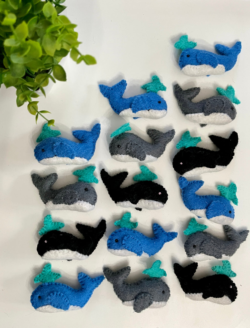 Multiple handmade felt whales in blue, gray, and black with turquoise water spouts arranged on white surface near green plant leaves