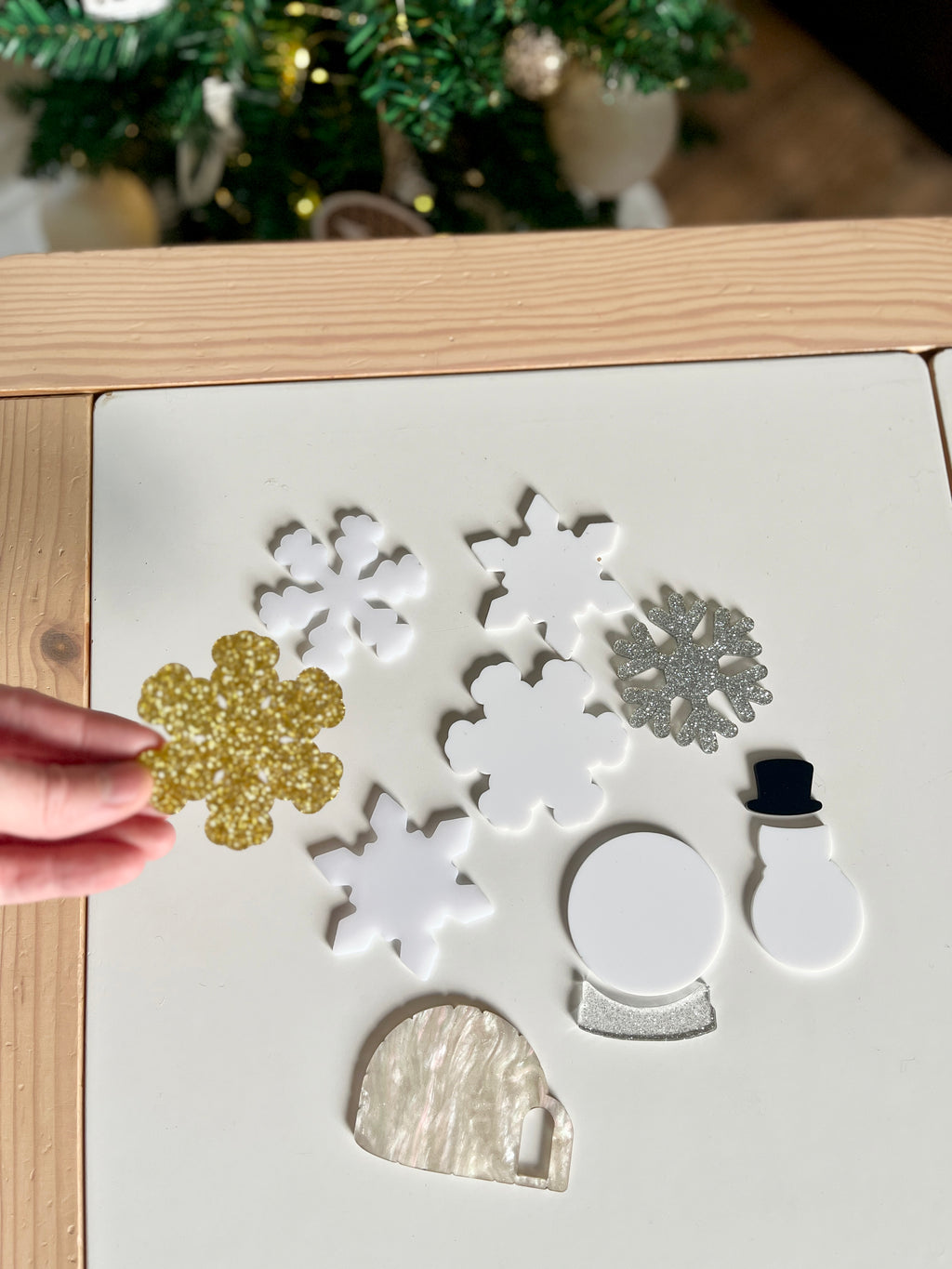 Hand holding gold glitter snowflake among white and silver acrylic holiday shapes including snowman, snow globe, and igloo on table