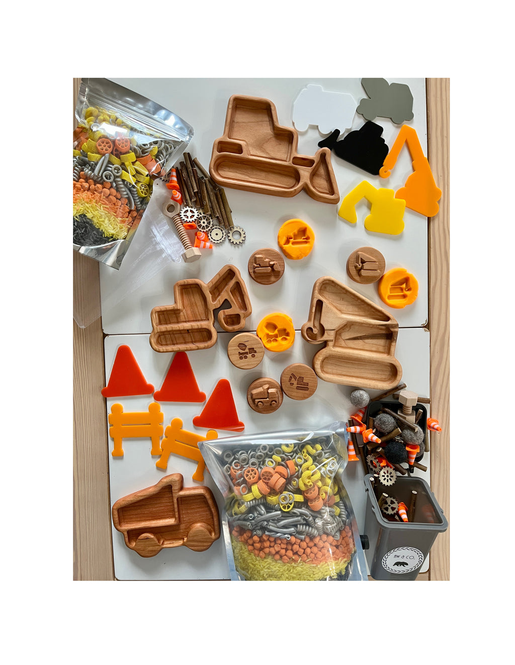 Wooden construction vehicle trays, round stamps with tools, plastic cones, benches, and bags of colorful craft pieces on white surface