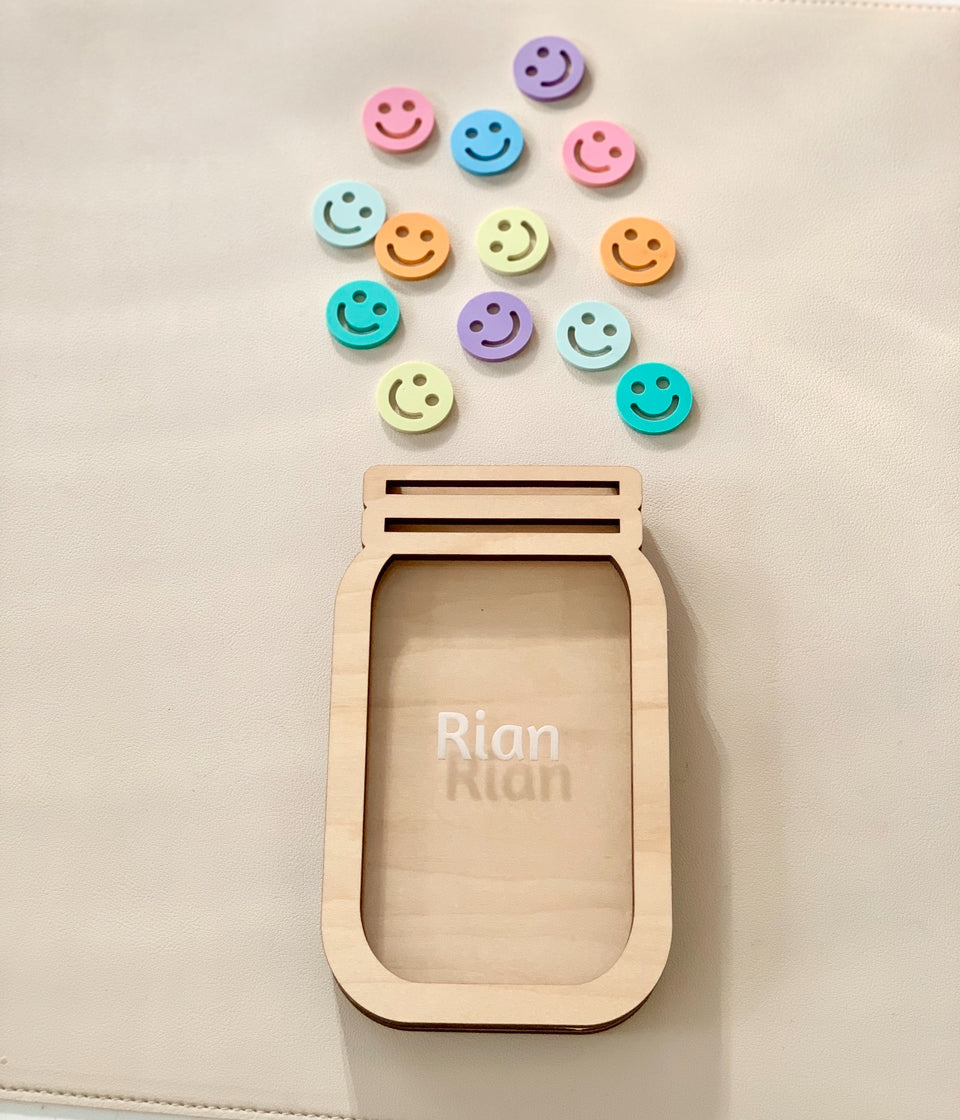 Light wooden jar frame engraved with the name Rian and colorful smiley face tokens arranged above on beige surface