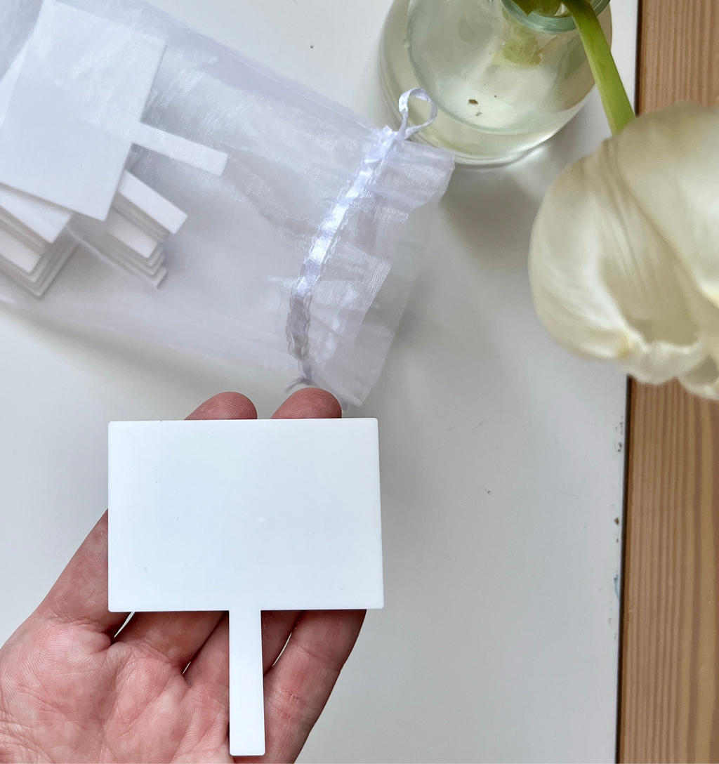 Hand holding a blank white plastic garden label with more stacked labels and a glass vase with white flowers nearby