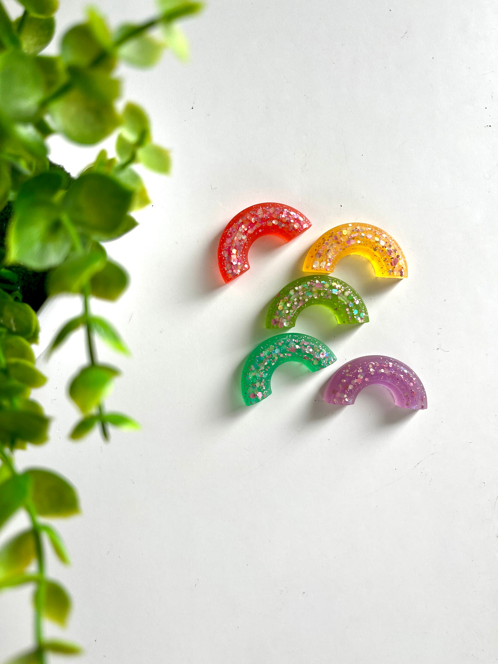 Five sparkly rainbow-shaped resin pieces in red, yellow, green, teal, and purple near green plant leaves on a white surface