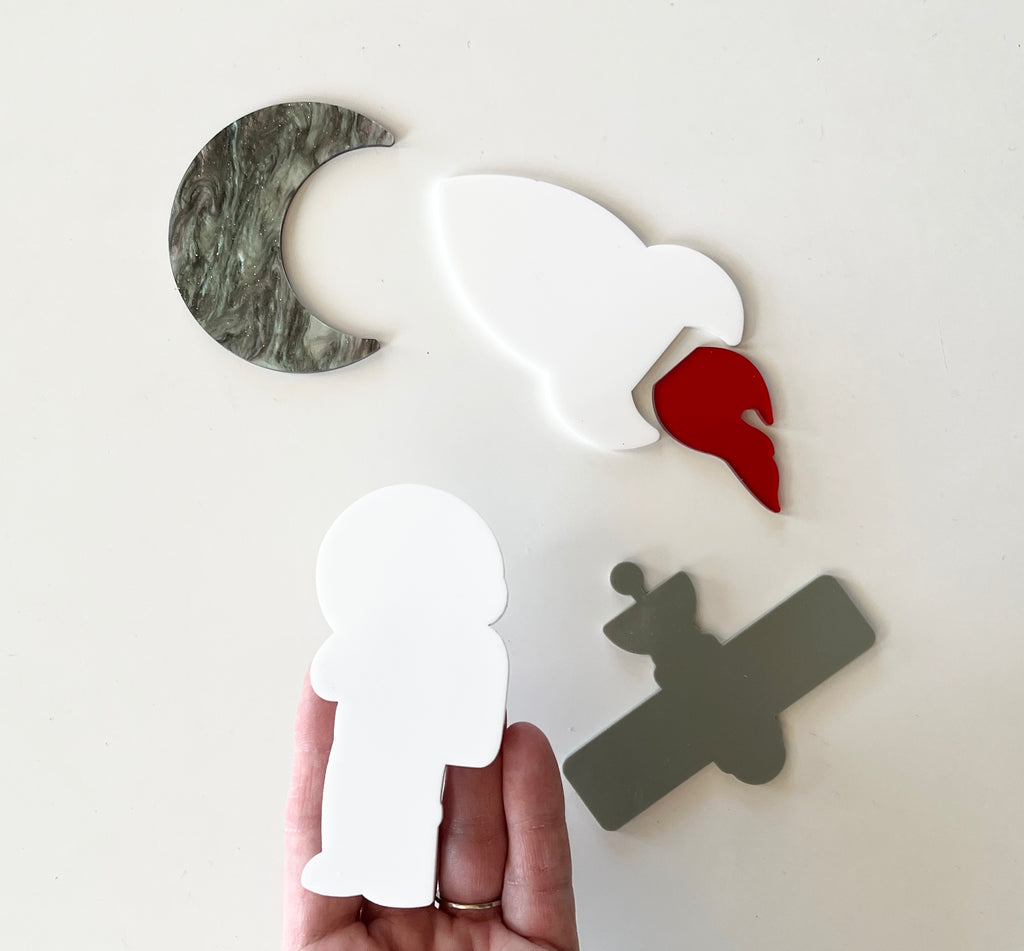 Hand holding white astronaut shape with green marble crescent moon, white bird with red detail, and gray satellite on white surface
