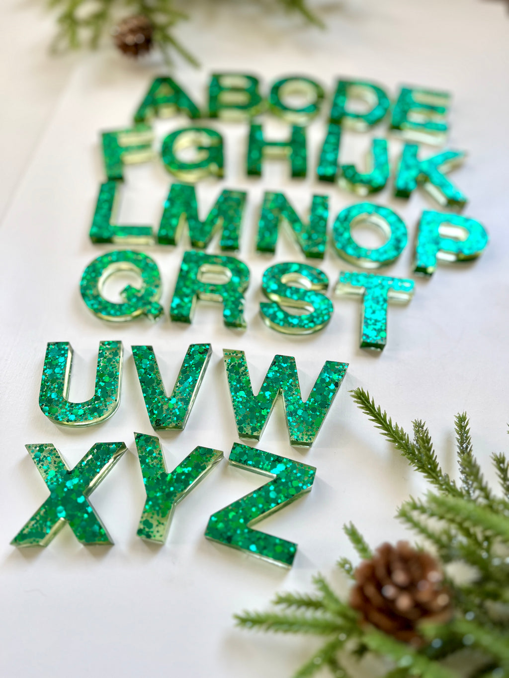 Green glitter resin alphabet letters arranged on white background with pine branches and pine cones nearby