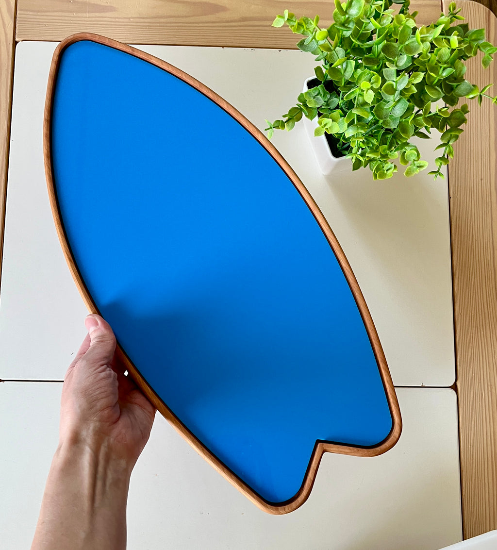 Hand holding a blue surfboard-shaped wooden tray above a light wooden table with a small green potted plant nearby