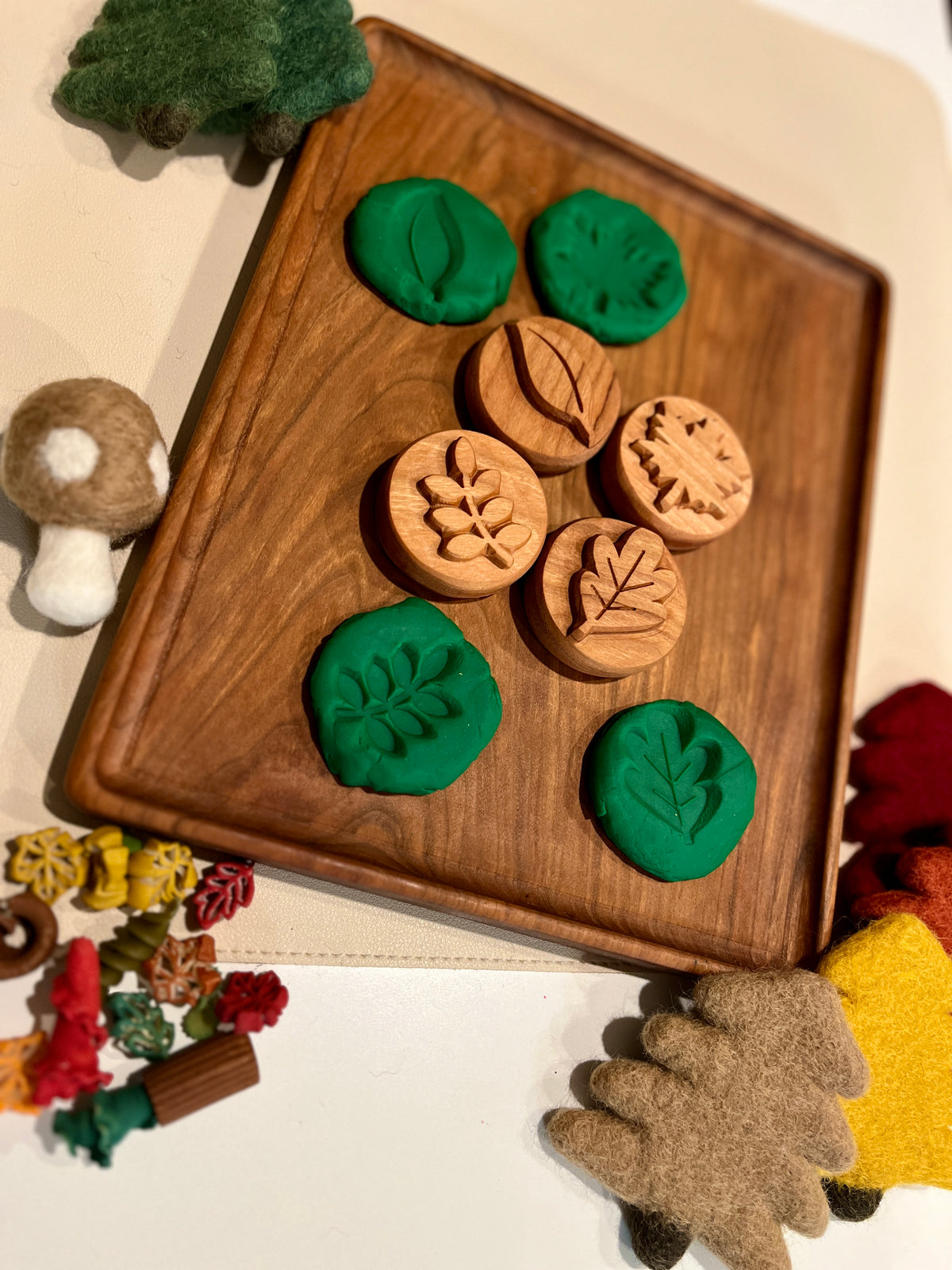 Wooden leaf-themed stamps with green playdough impressions on a wooden tray surrounded by felt mushrooms and autumn decorations