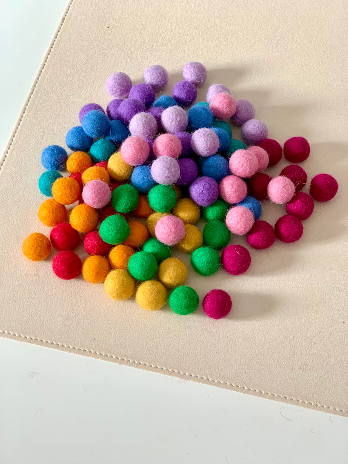 Pile of colorful 1.5cm felt balls in rainbow shades scattered on light beige surface