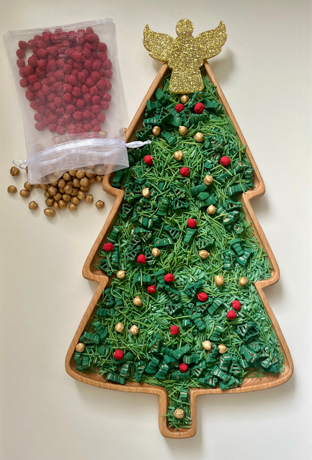 Christmas tree-shaped wooden tray filled with green shredded paper, red and gold decorative beads, and topped with a glittery gold angel