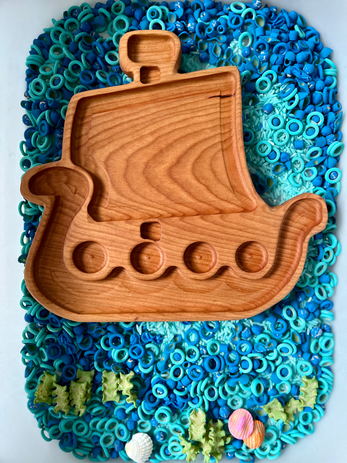 Wooden Viking ship-shaped tray with carved compartments resting on blue and green sensory materials with shells at the bottom