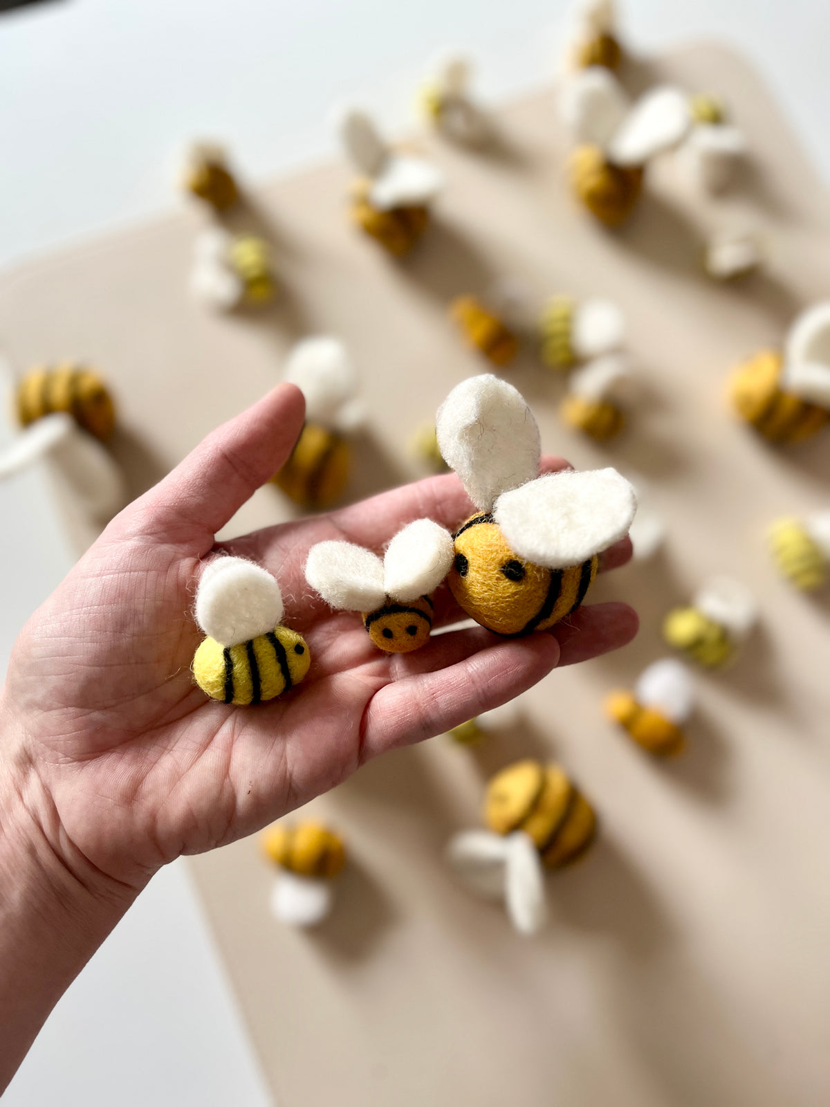Hand holding three felt bees of varying sizes with yellow and black stripes and white wings on a beige surface with more bees scattered
