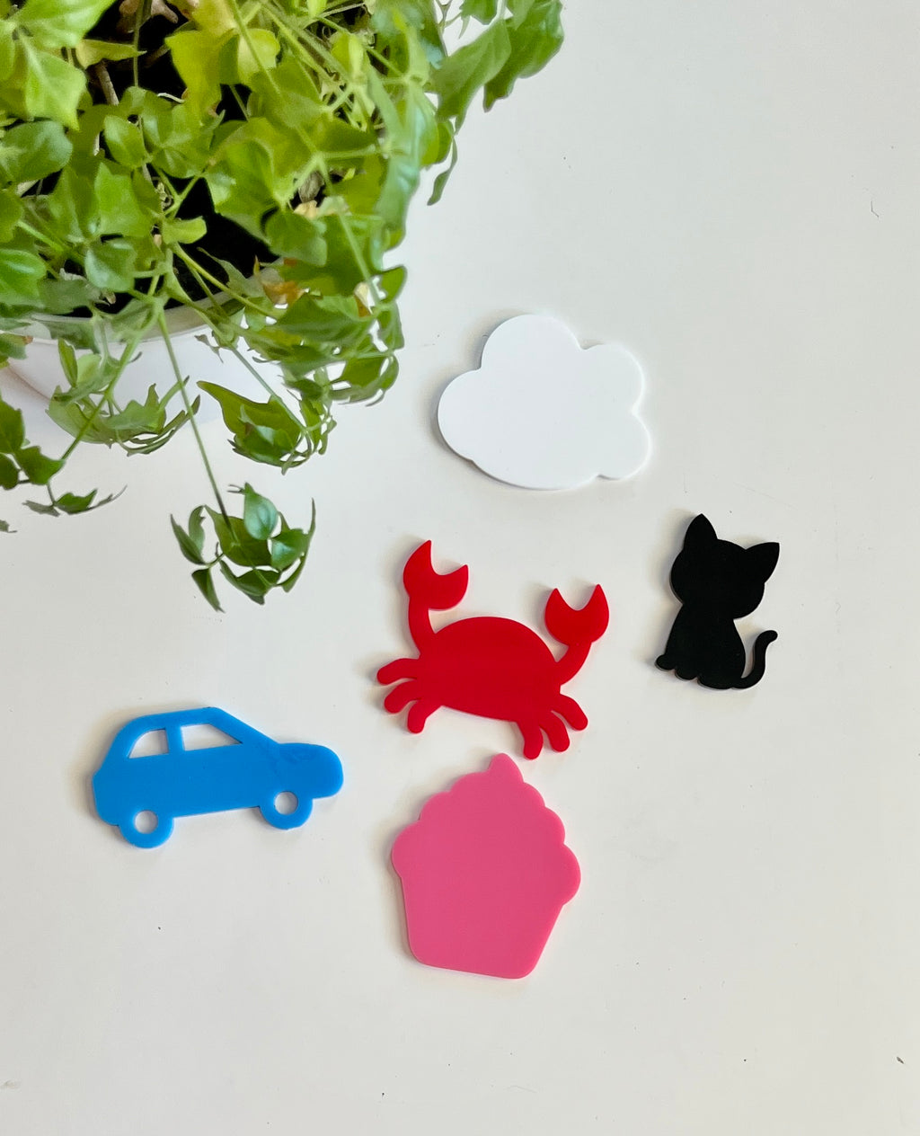 Five colorful acrylic shapes including a white cloud, red crab, black cat, blue car, and pink cupcake on white surface with green plant nearby