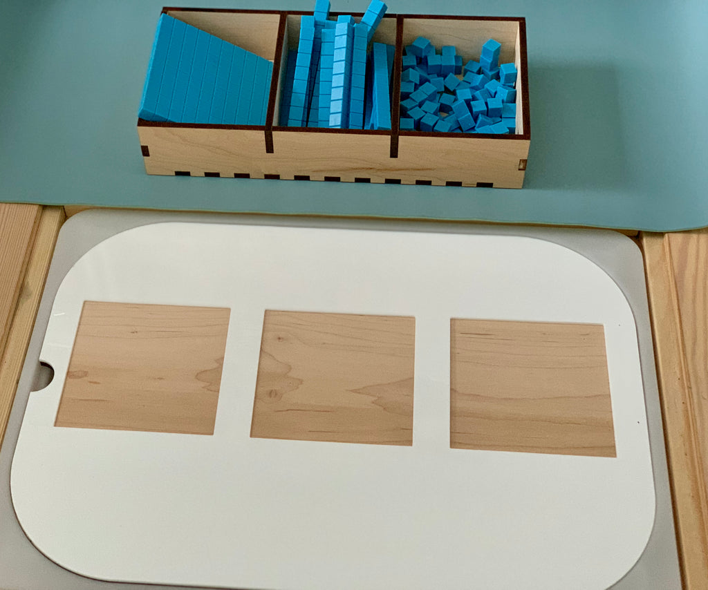 Wooden box holding blue base ten blocks divided into hundreds, tens, and ones above a white board with three wooden square cutouts