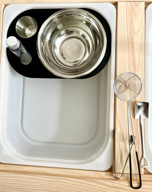 White tray holding stainless steel mixing bowls, a small bottle, and a metal cup beside wooden utensils on a light wood surface