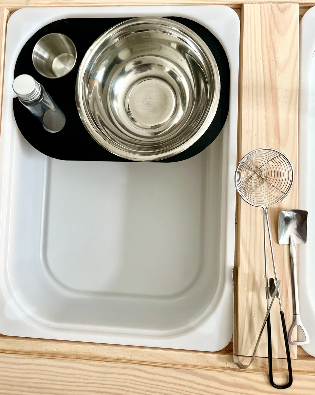 White tray holding stainless steel mixing bowls, a small bottle, and a metal cup beside wooden utensils on a light wood surface