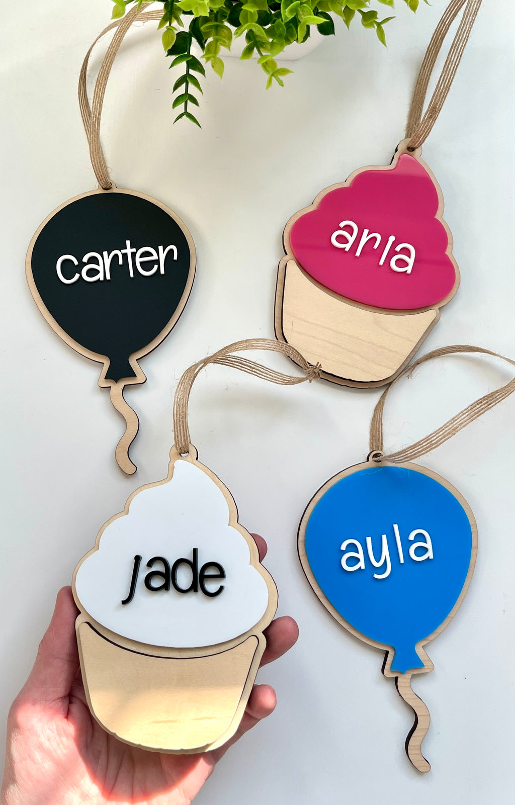 Four wooden hanging tags shaped like balloons and cupcakes in black, blue, pink, and white with personalized letters