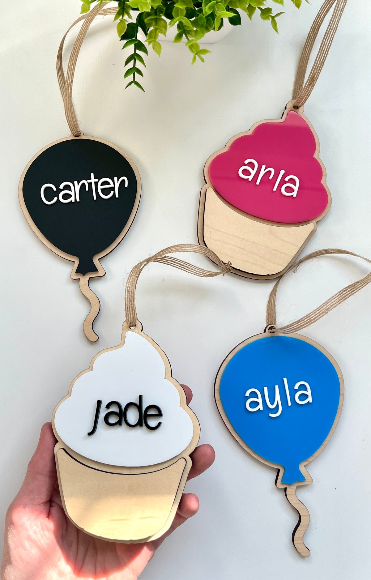 Four wooden hanging tags shaped like balloons and cupcakes in black, blue, pink, and white with personalized letters
