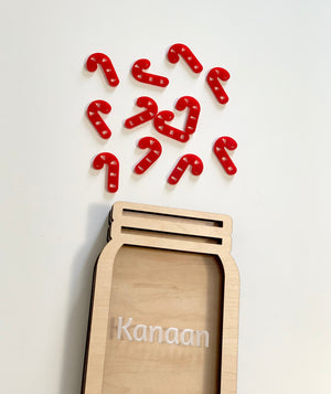 Natural wooden jar-shaped frame personalized with the name Kanaan and scattered red candy cane tokens on white background