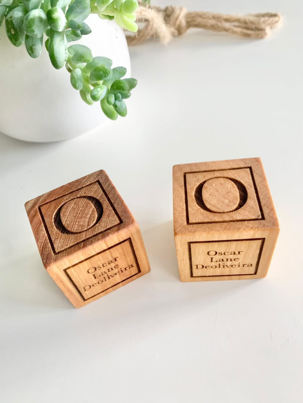 Two personalized wooden baptism blocks with engraved names on white surface next to green succulent plant