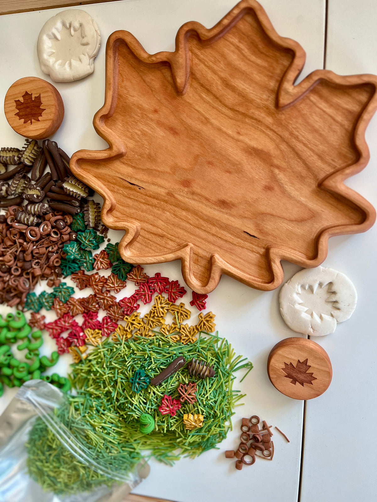 Leaf-shaped wooden tray with colorful sensory materials, leaf stamps, and white clay impressions on a white surface.