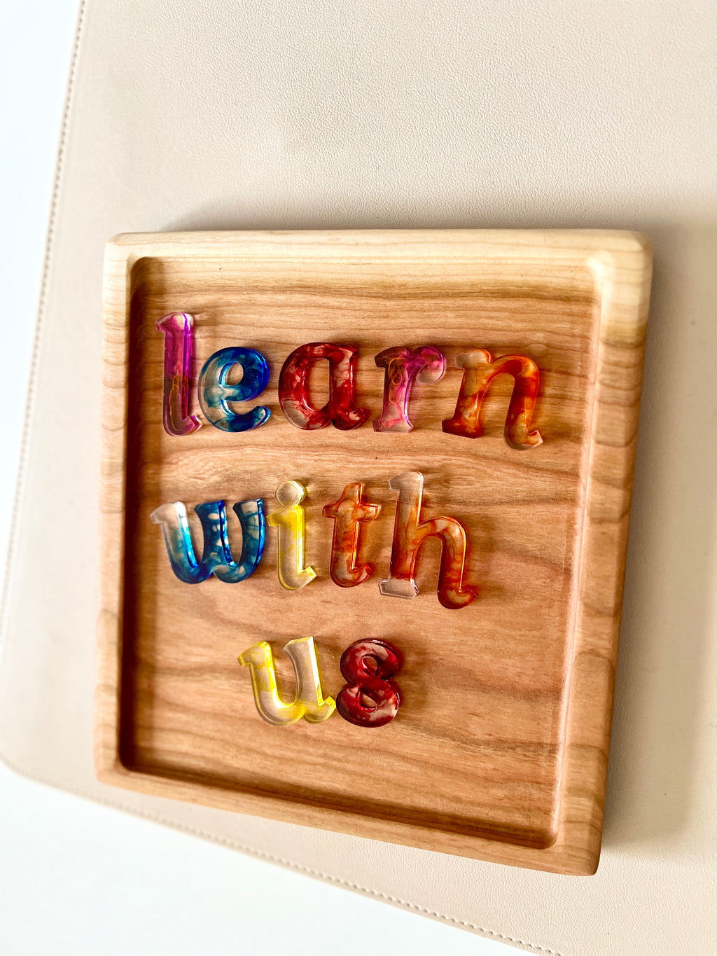 Wooden tray with multicolored translucent lowercase letters spelling "learn with us" on beige surface