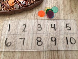 Clear plastic board with numbers one to ten written in separate squares on a light wood surface with colorful discs nearby