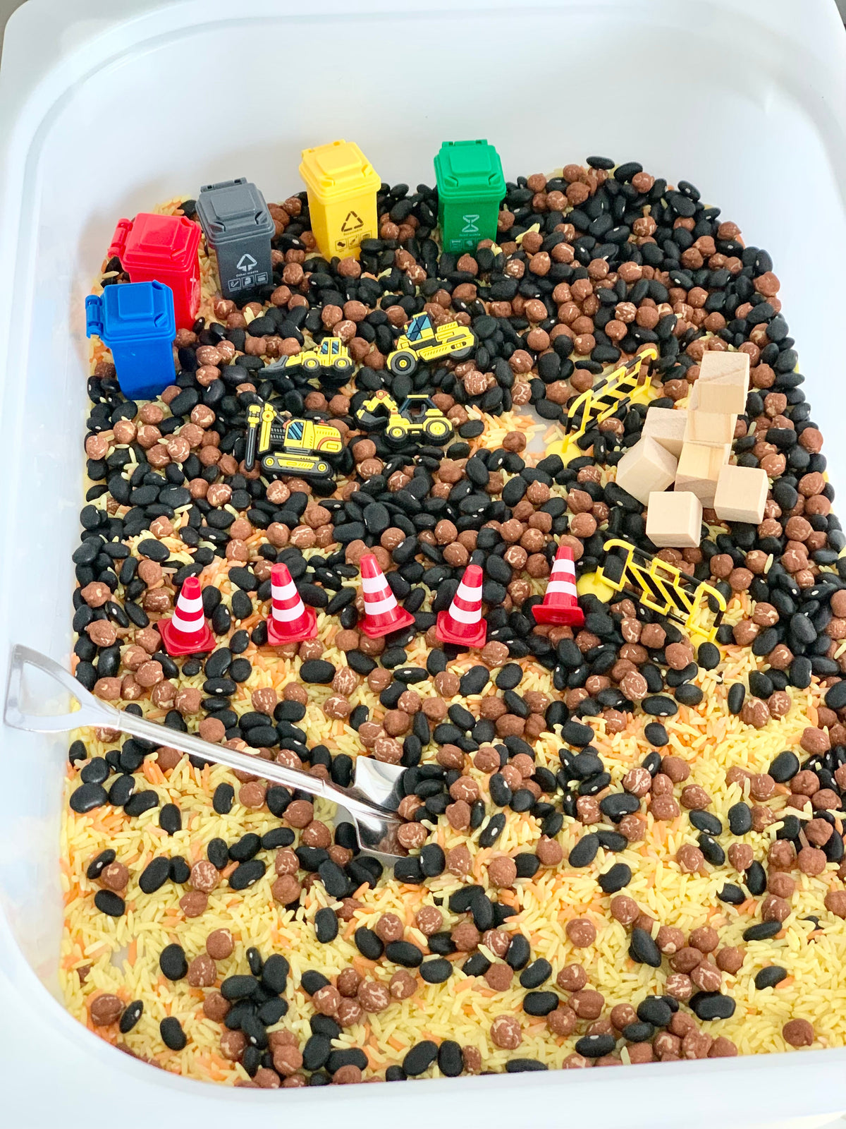 Miniature construction vehicles, traffic cones, and colorful recycling bins arranged on mixed beans and rice in a sensory bin