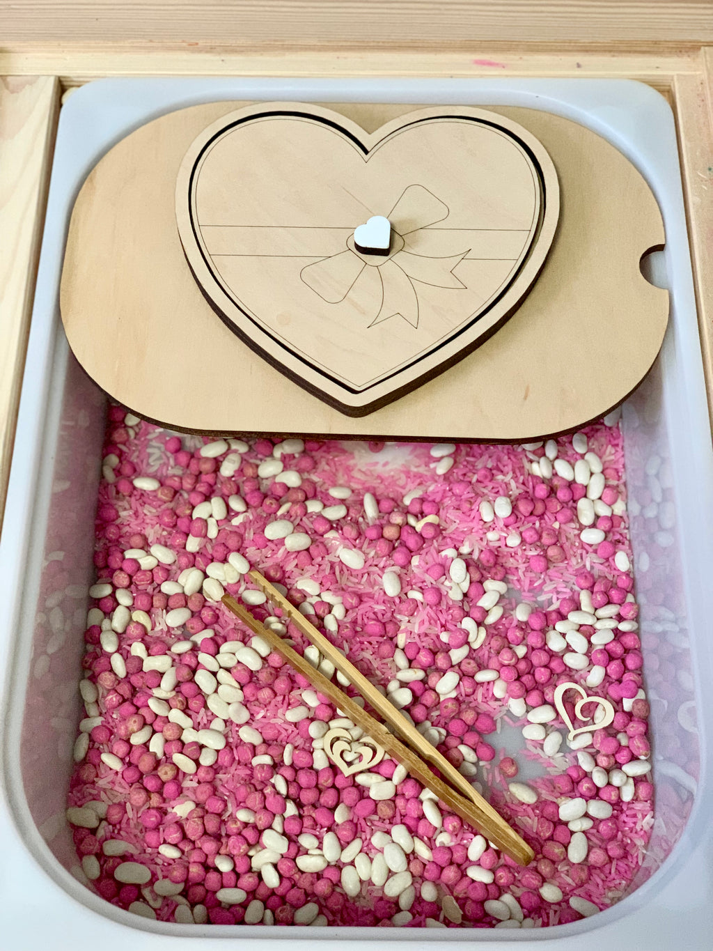 Light wooden heart-shaped lid with bow engraving over a bin filled with pink and white sensory beans and wooden tweezers